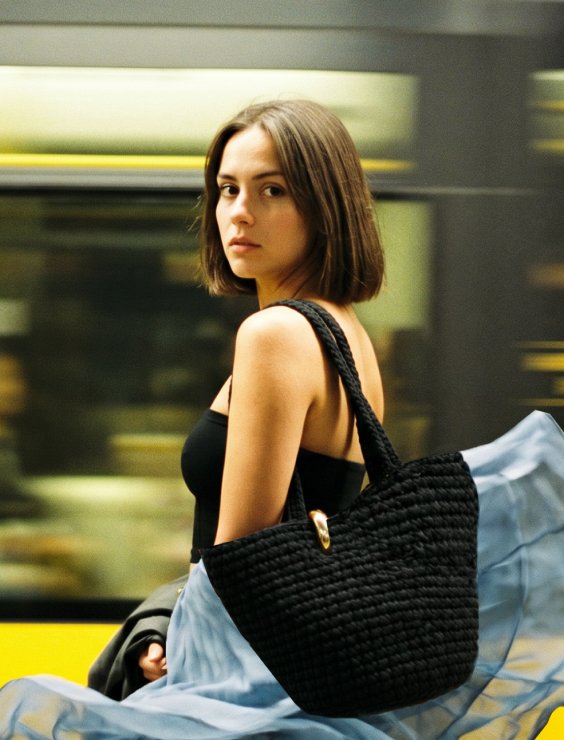 Portait d'une femme portant un sac d'épaule noir fait main dans un décor urbain flou.